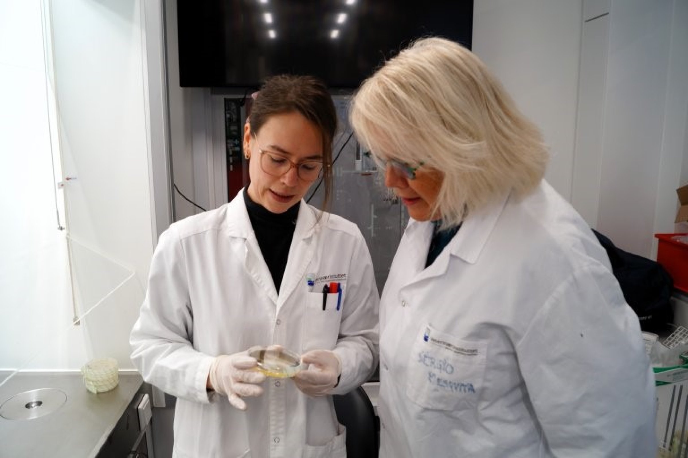 Forskar Ida Skaar og masterstudenten hennar, Marte Mølmann Kåråsen, står i eit laboratorium i hvite frakkar og ser sammen på ein petriskål.